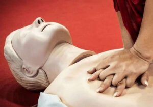 A person performs chest compressions on a CPR training mannequin with white nails and a red background during a practical onsite or group training session.