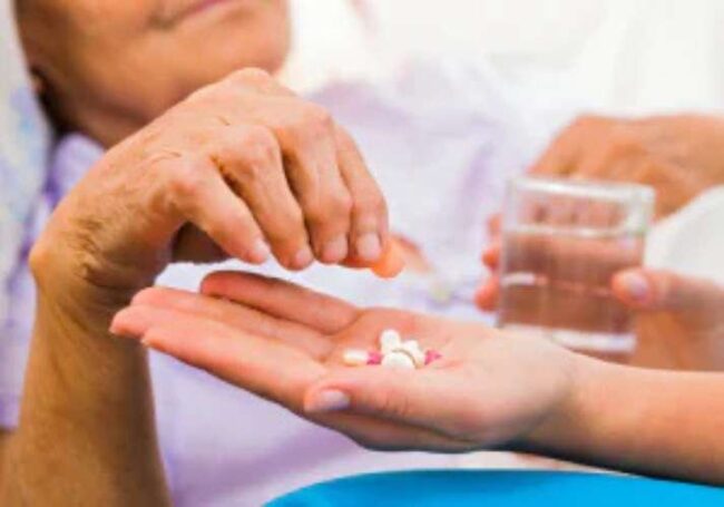 A close-up image showing one person's hands offering several pills and capsules to another person's hand, who is also holding a glass of water, illustrating medication administration.